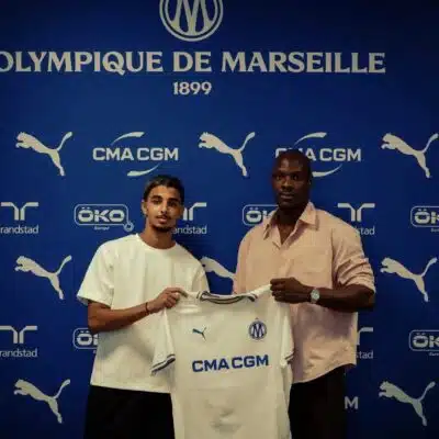 Two men pose with a white CMA CGM football jersey against a blue Olympique de Marseille backdrop with sponsor logos nearby, indoors.
