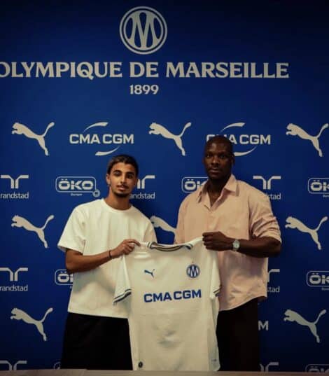 Two men pose with a white CMA CGM football jersey against a blue Olympique de Marseille backdrop with sponsor logos nearby, indoors.