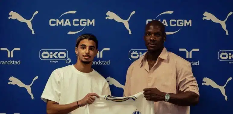 Two men pose with a white CMA CGM football jersey against a blue Olympique de Marseille backdrop with sponsor logos nearby, indoors.