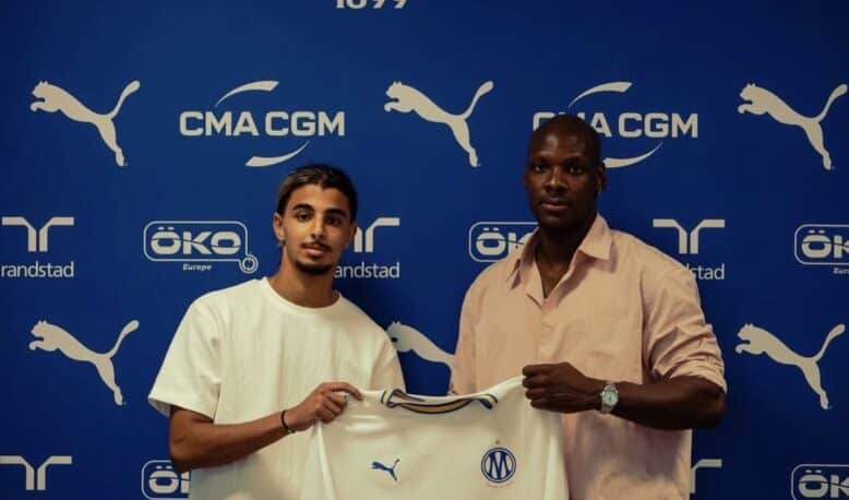 Two men pose with a white CMA CGM football jersey against a blue Olympique de Marseille backdrop with sponsor logos nearby, indoors.