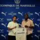 Two men pose with a white CMA CGM football jersey against a blue Olympique de Marseille backdrop with sponsor logos nearby, indoors.