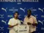 Two men pose with a white CMA CGM football jersey against a blue Olympique de Marseille backdrop with sponsor logos nearby, indoors.