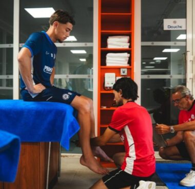 Man in blue shirt sits on a table while two therapists in red shirts treat his foot in a clinic setting.