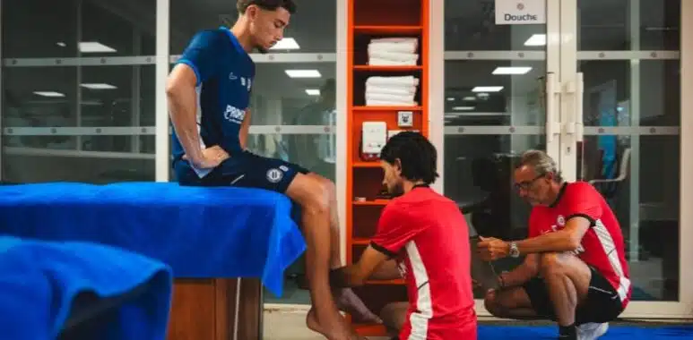 Man in blue shirt sits on a table while two therapists in red shirts treat his foot in a clinic setting.