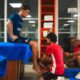 Man in blue shirt sits on a table while two therapists in red shirts treat his foot in a clinic setting.