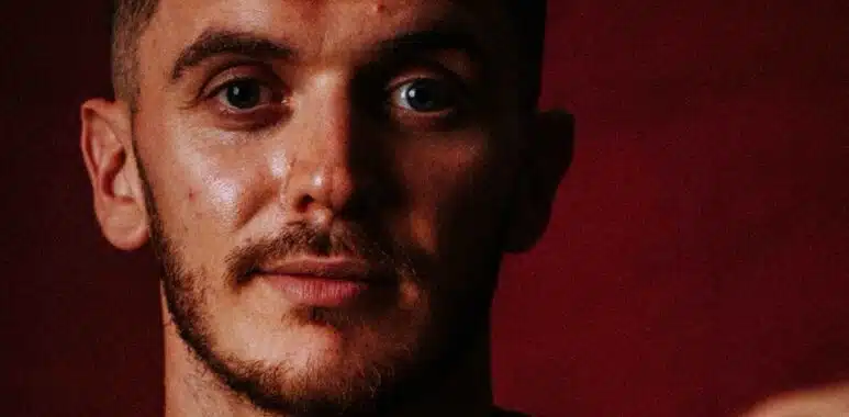 Close-up of a man with stubble and short hair, against a dark red background.
