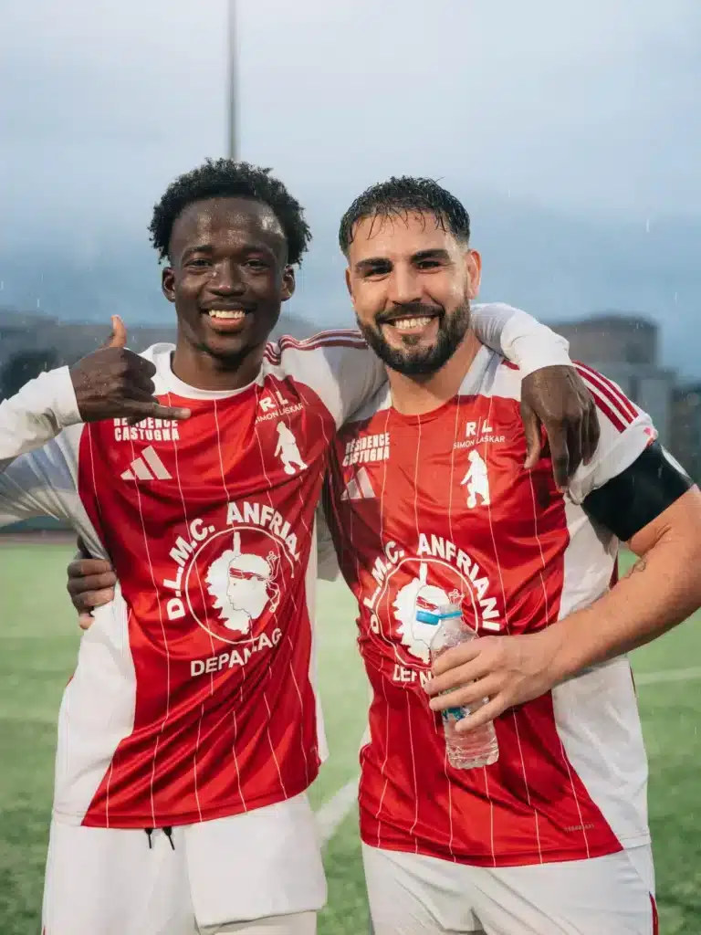 Two male soccer players in red uniforms smile and hug on a rainy field, one holding a water bottle and making a friendly gesture with his hand on the other's shoulder