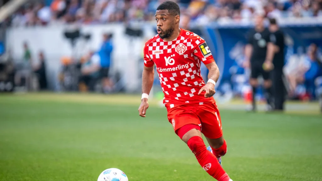 Soccer player in a red checkered jersey dribbles the ball on a green field with a blurred stadium crowd in the background.