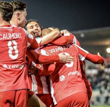 Group of soccer players in red uniforms celebrate with hugs on the field at night, stadium lights glowing in the background.
