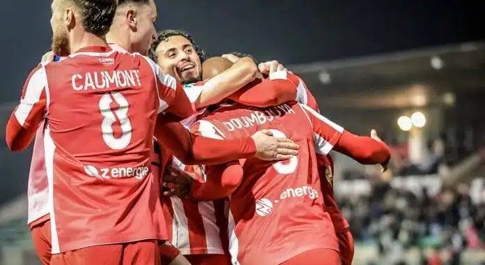 as cannes | AllezPaillade.com Group of soccer players in red uniforms celebrate with hugs on the field at night, stadium lights glowing in the background.