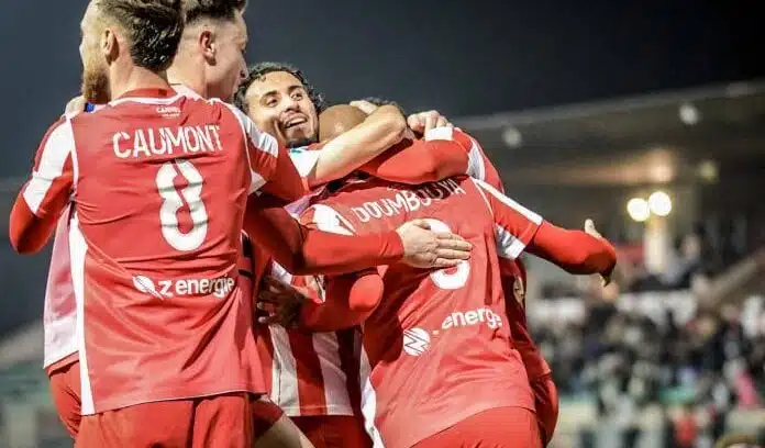 Group of soccer players in red uniforms celebrate with hugs on the field at night, stadium lights glowing in the background.