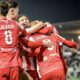 Group of soccer players in red uniforms celebrate with hugs on the field at night, stadium lights glowing in the background.