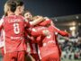 Group of soccer players in red uniforms celebrate with hugs on the field at night, stadium lights glowing in the background.