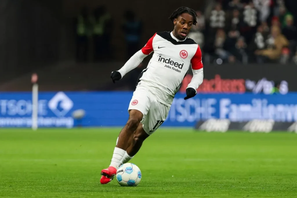 Soccer player in a white Eintracht Frankfurt kit with red sleeves dribbles the ball on a green pitch during a match, sponsor logo visible on jersey.