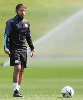Male soccer player in black training gear standing on a sunny green field with a ball at his feet