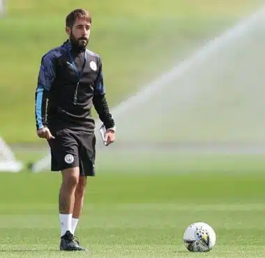 Male soccer player in black training gear standing on a sunny green field with a ball at his feet