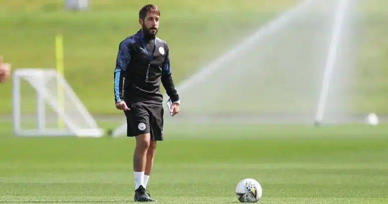 Male soccer player in black training gear standing on a sunny green field with a ball at his feet