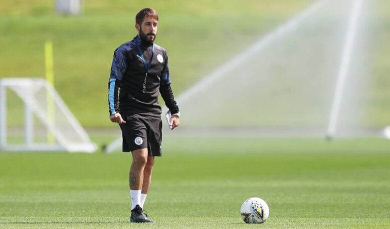 Male soccer player in black training gear standing on a sunny green field with a ball at his feet