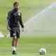 Male soccer player in black training gear standing on a sunny green field with a ball at his feet