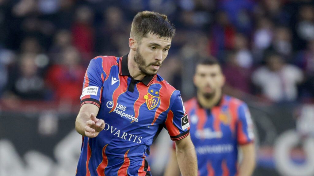 BASEL - YB | AllezPaillade.com Soccer player in a blue and red striped kit reaching forward during a match with a blurred stadium crowd behind him.