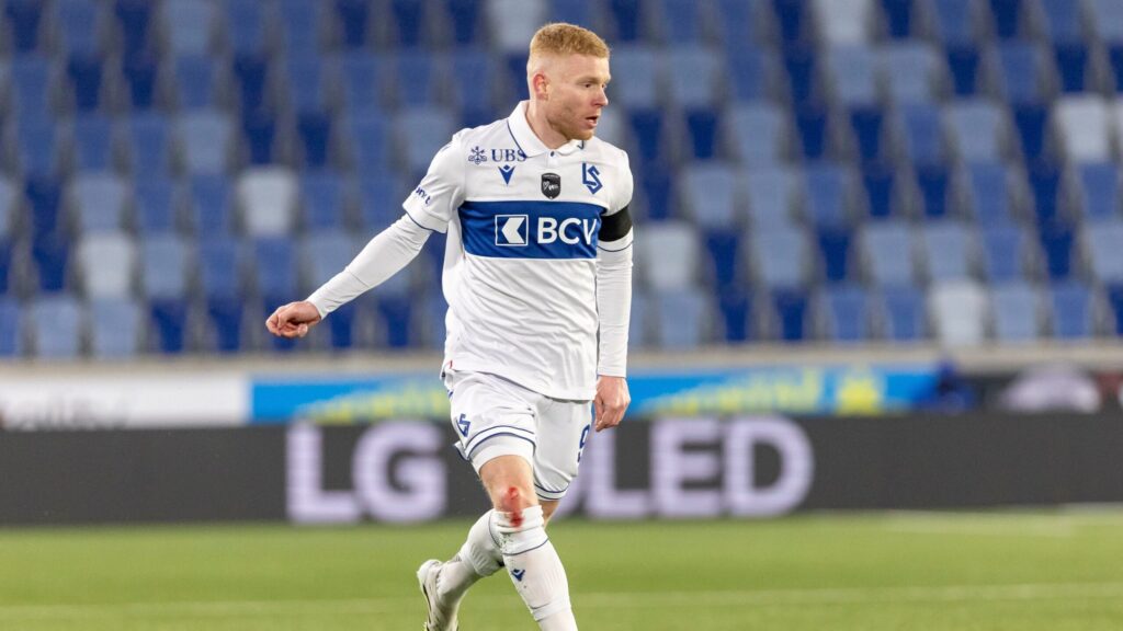 mollet lausanne | AllezPaillade.com Soccer player in a white kit with a blue chest stripe sprinting on a green pitch, stadium seats in the background