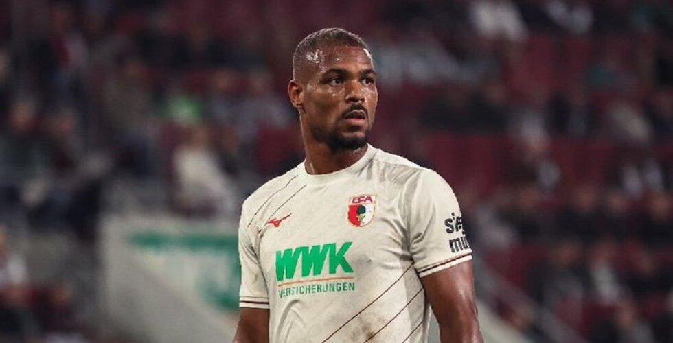 Footballer in a white jersey on a stadium pitch with a blurred crowd in the background.