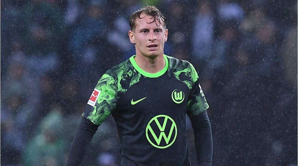 Male soccer player in a black and green Wolfsburg jersey stands on the pitch in rain, looking focused with hands at sides.