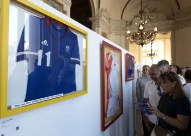 Framed sports jerseys on yellow-trimmed display panels in a grand, ornate room with visitors reading brochures.