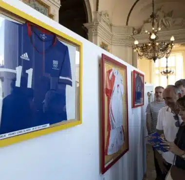 Framed sports jerseys on yellow-trimmed display panels in a grand, ornate room with visitors reading brochures.