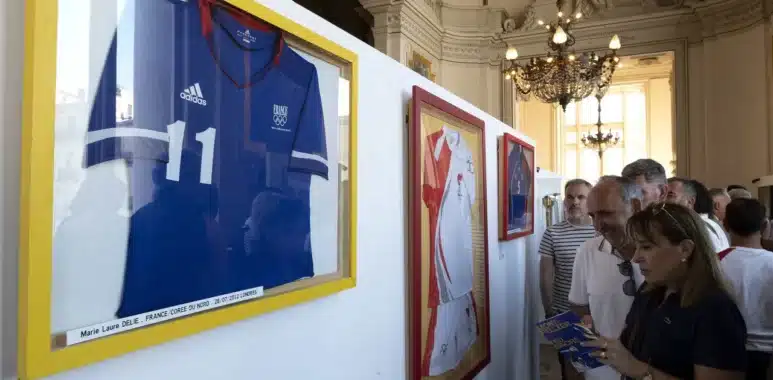 ollection-olympique-ouis-icollin | AllezPaillade.com Framed sports jerseys on yellow-trimmed display panels in a grand, ornate room with visitors reading brochures.