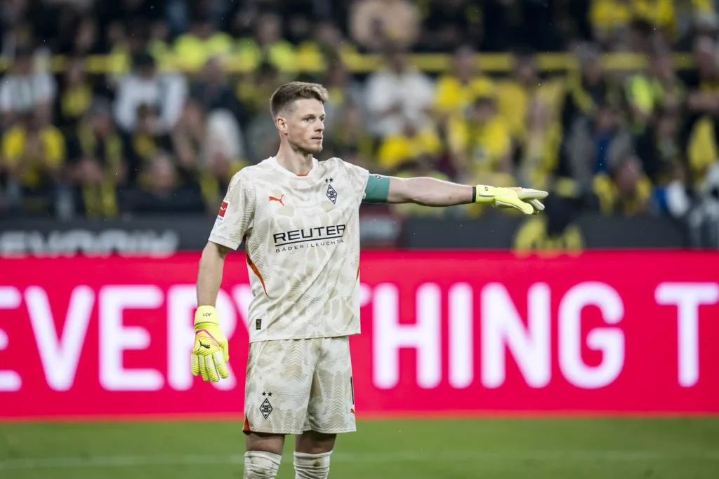 Borussia Dortmund goalkeeper in a white patterned kit with yellow gloves on a football pitch, extending his right arm toward the stands.