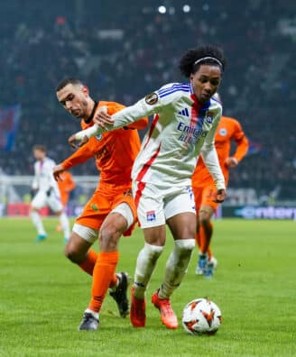 Soccer players chase the ball on a grassy field during a match, one in a white jersey with blue stripes and red accents, the other in orange, stadium crowd in the background.
