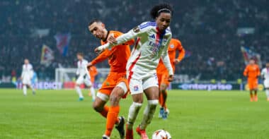Soccer players chase the ball on a grassy field during a match, one in a white jersey with blue stripes and red accents, the other in orange, stadium crowd in the background.