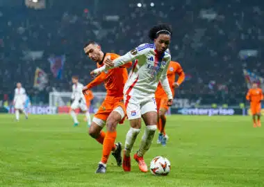 Soccer players chase the ball on a grassy field during a match, one in a white jersey with blue stripes and red accents, the other in orange, stadium crowd in the background.