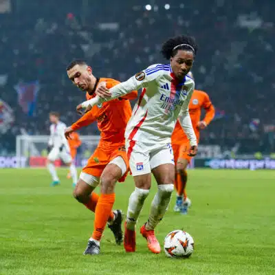 Soccer players chase the ball on a grassy field during a match, one in a white jersey with blue stripes and red accents, the other in orange, stadium crowd in the background.