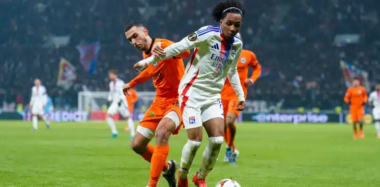 Soccer players chase the ball on a grassy field during a match, one in a white jersey with blue stripes and red accents, the other in orange, stadium crowd in the background.