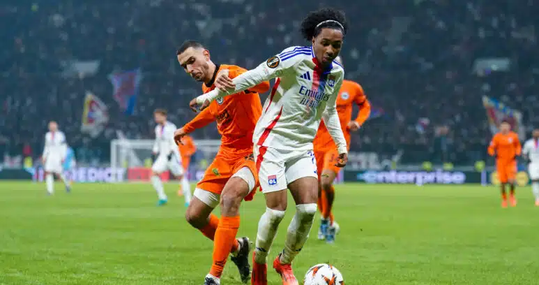 Soccer players chase the ball on a grassy field during a match, one in a white jersey with blue stripes and red accents, the other in orange, stadium crowd in the background.