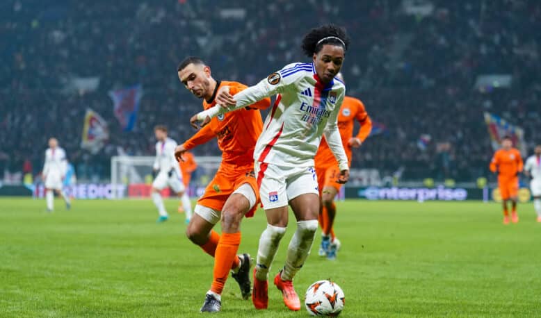 Soccer players chase the ball on a grassy field during a match, one in a white jersey with blue stripes and red accents, the other in orange, stadium crowd in the background.