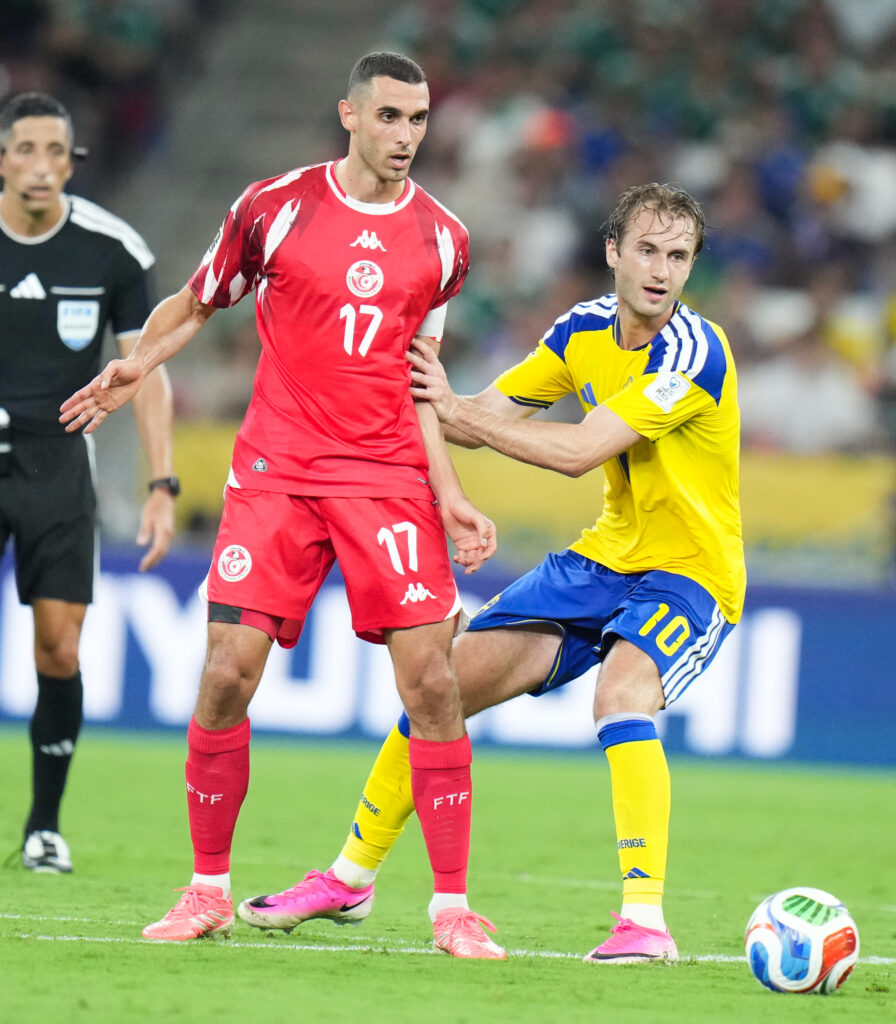 Two soccer players contest the ball on a green pitch; one in a red jersey (#17) and the other in yellow and blue reach for it from the side.