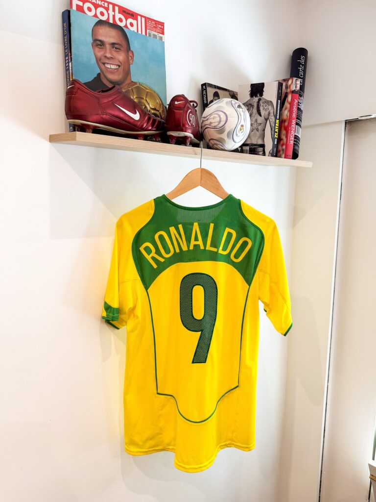 1784026692466 | AllezPaillade.com Yellow Ronaldo soccer jersey with green trim hanging on a wooden hanger beneath a shelf displaying football boots, a ball, and magazines.