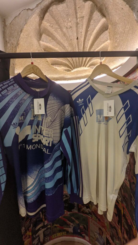 1876c38d-34b6-4f6e-a7ca-18b8e412e7a6 | AllezPaillade.com Two Adidas jerseys on wooden hangers hanging from a metal rod in a shop display, with a carved stone circular arch in the background.