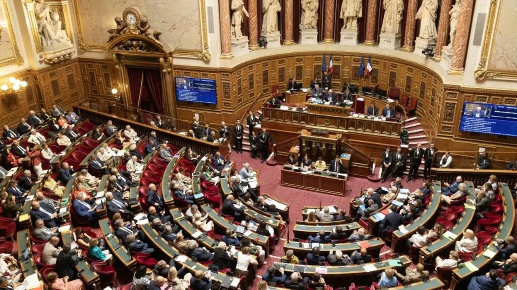 6a5f78e27712a217521758 | AllezPaillade.com Parliament chamber with curved rows of red seats, officials, and a central dais where speakers address the assembly.