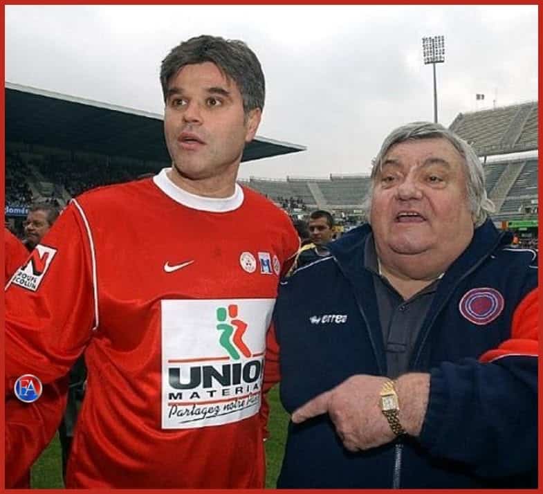 723792782_1318011243825064_6508185216415394543_n | AllezPaillade.com Two men pose on a soccer field; one in a red jersey with UNIO MATERIA sponsor, the other in a dark jacket, with a stadium in the background.