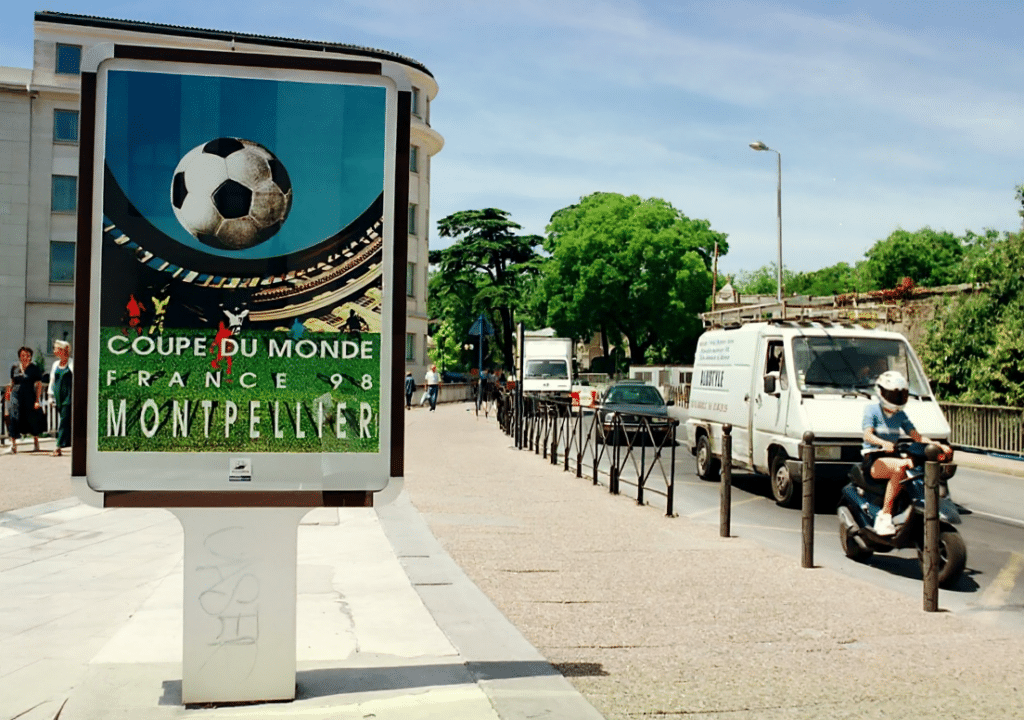 73dca0c9-6f98-4ce6-a093-3b3dc20586f5 | AllezPaillade.com Poster advertising the 1998 World Cup in Montpellier, showing a soccer ball and the text 'COUPE DU MONDE FRANCE 98 MONTPELLIER' on a blue background beside the sidewalk.