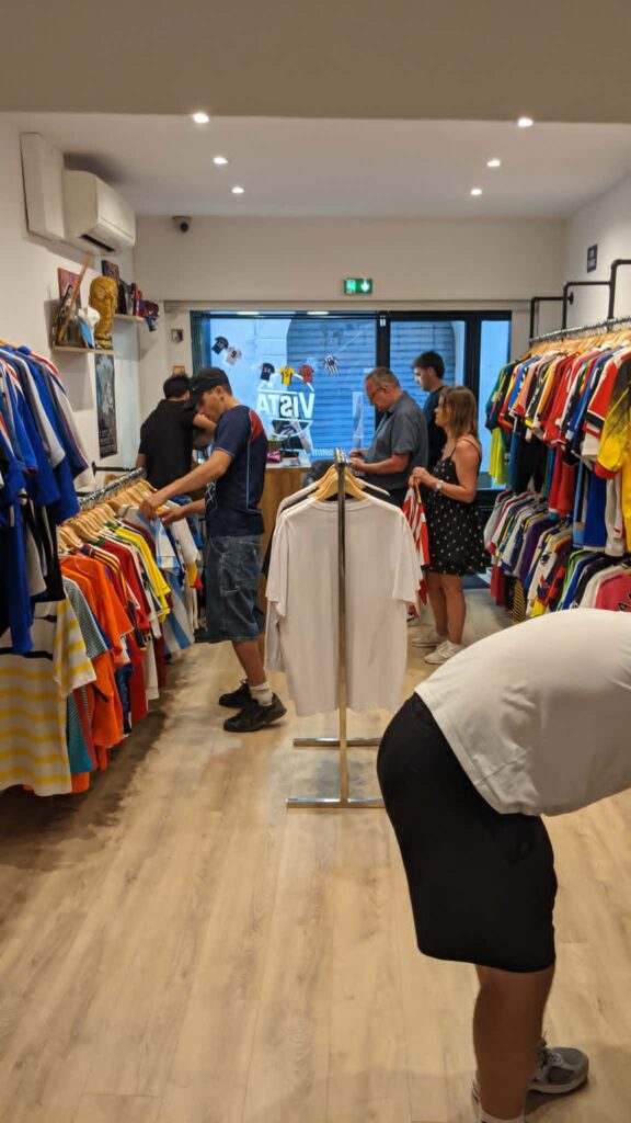 74bafcb6-d6d3-4333-8d81-32a920d01975 | AllezPaillade.com Inside a clothing store with racks of colorful shirts on both sides; customers browsing."Most important info: shopping scene in a apparel store"