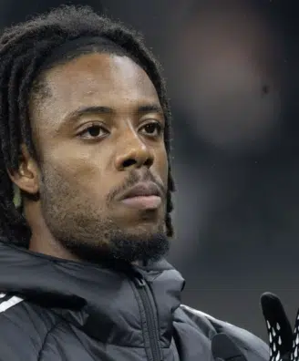 Close-up of a Black man with dreadlocks wearing a dark jacket and gloves, looking upward.