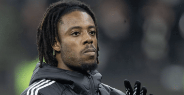 Close-up of a Black man with dreadlocks wearing a dark jacket and gloves, looking upward.