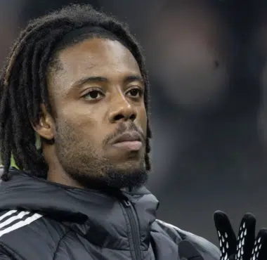 Close-up of a Black man with dreadlocks wearing a dark jacket and gloves, looking upward.