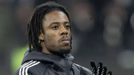 Close-up of a Black man with dreadlocks wearing a dark jacket and gloves, looking upward.