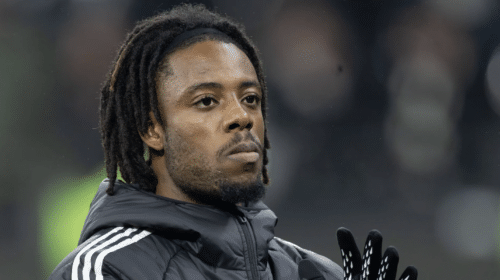 Close-up of a Black man with dreadlocks wearing a dark jacket and gloves, looking upward.
